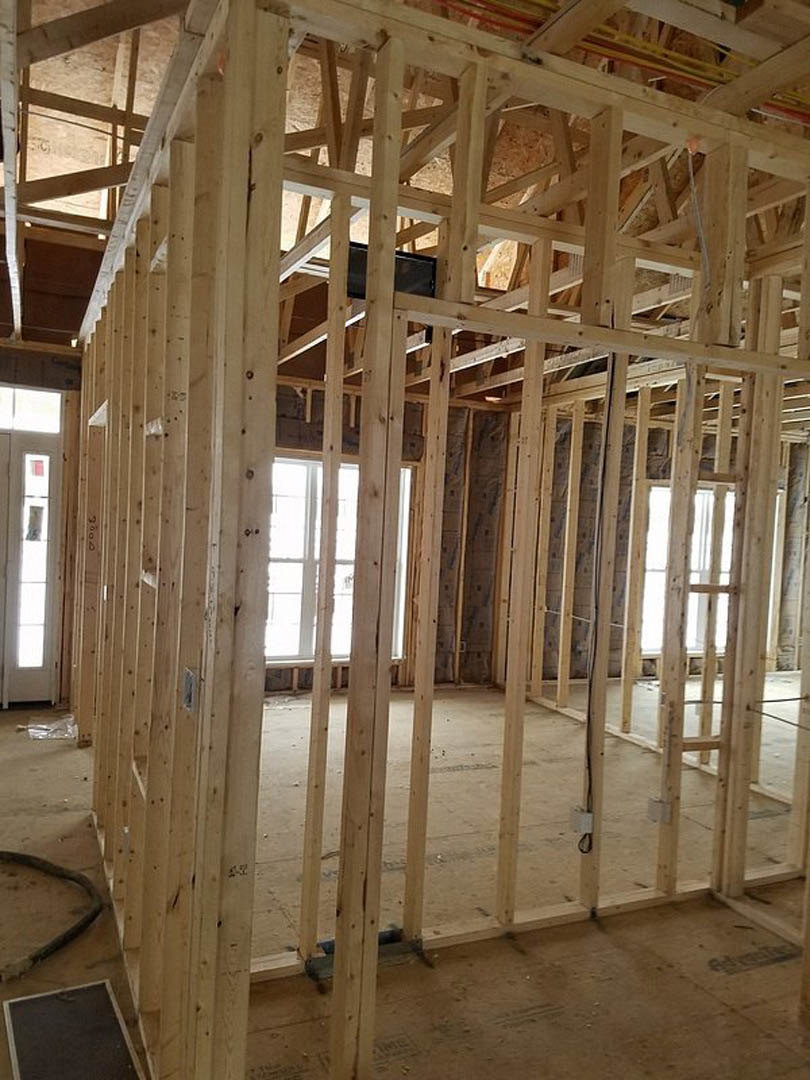 Exposed wood framing and beams with construction materials, white-framed window letting in natural light, black square object resting on subfloor, hose visible in foreground