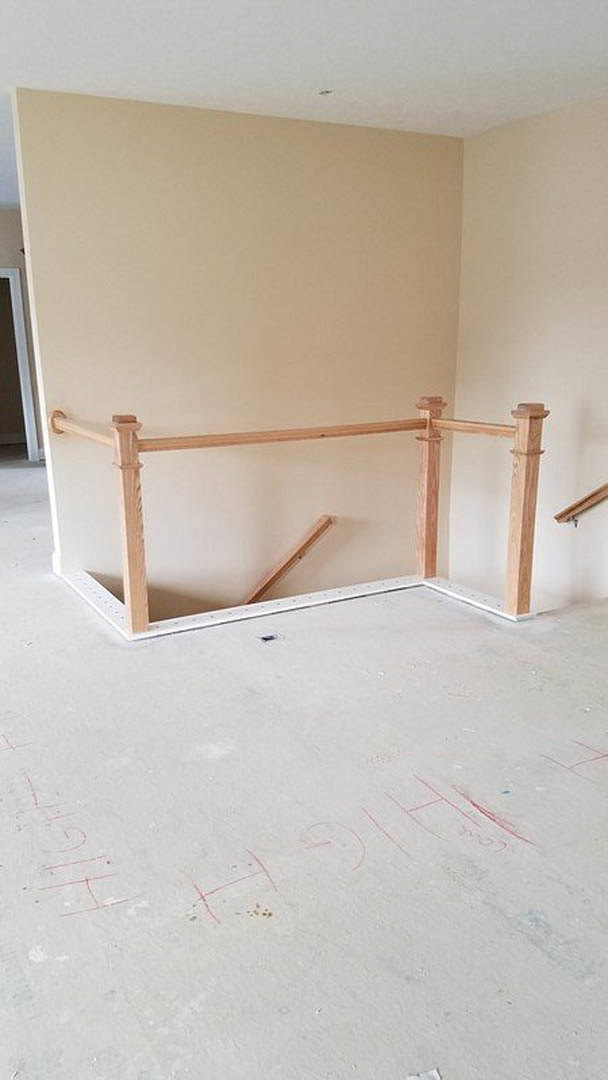 Wooden staircase with vertical railing, white plaster walls, light wood flooring featuring red markings, and close-up of a wooden post adjacent to a framed wall.