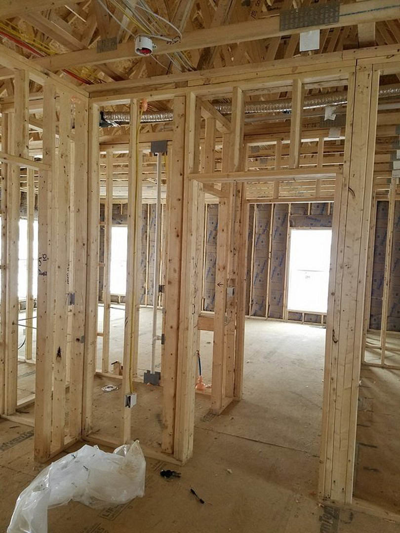 Exposed wood framing and beams in a house under construction, with unfinished walls, ceiling, and doorway; cardboard box with plastic bag, white door with black trim, white paper