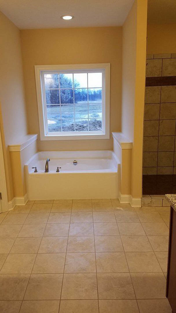 White freestanding tub beneath a large window, chrome faucet, light gray tile flooring, wood vanity cabinetry, and neutral tiled walls