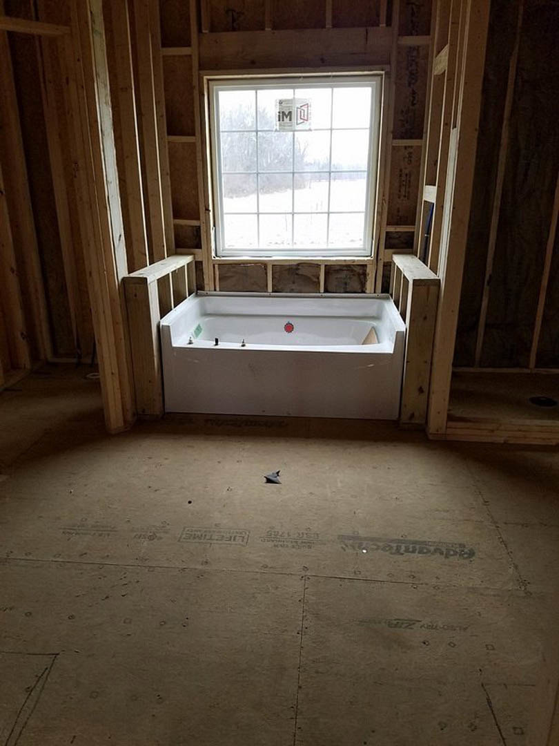 White freestanding bathtub with red emblem sits on unfinished concrete floor, window with construction sign in background, exposed walls and plumbing hole visible.