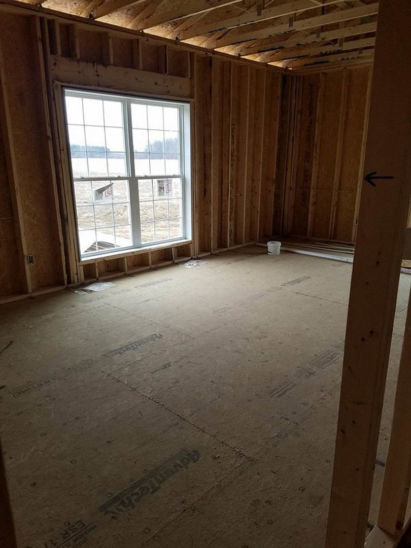 Sunlit room with unfinished wood flooring, exposed wooden beam, plastered wall, and large window overlooking outdoor dirt with visible tire tracks; white bucket near window.