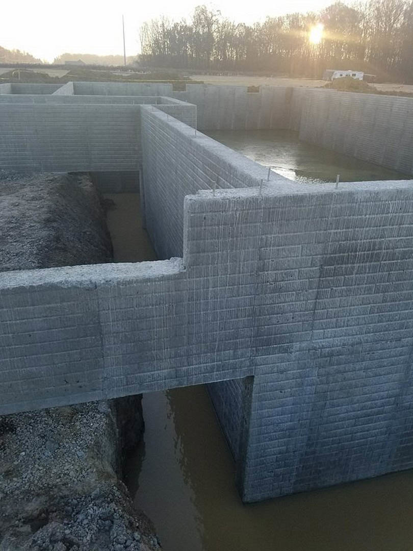 Concrete retaining wall filled with water, sunlight filtering through nearby trees, rocky slope visible in foreground.