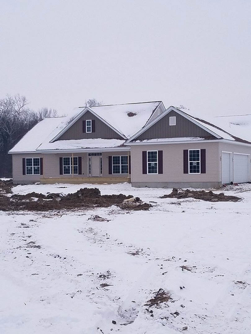 Two-story home with white siding and black trim, snow covering the roof and ground, white door with black handle, grid-style windows, rocks and a dog resting in the snowy yard