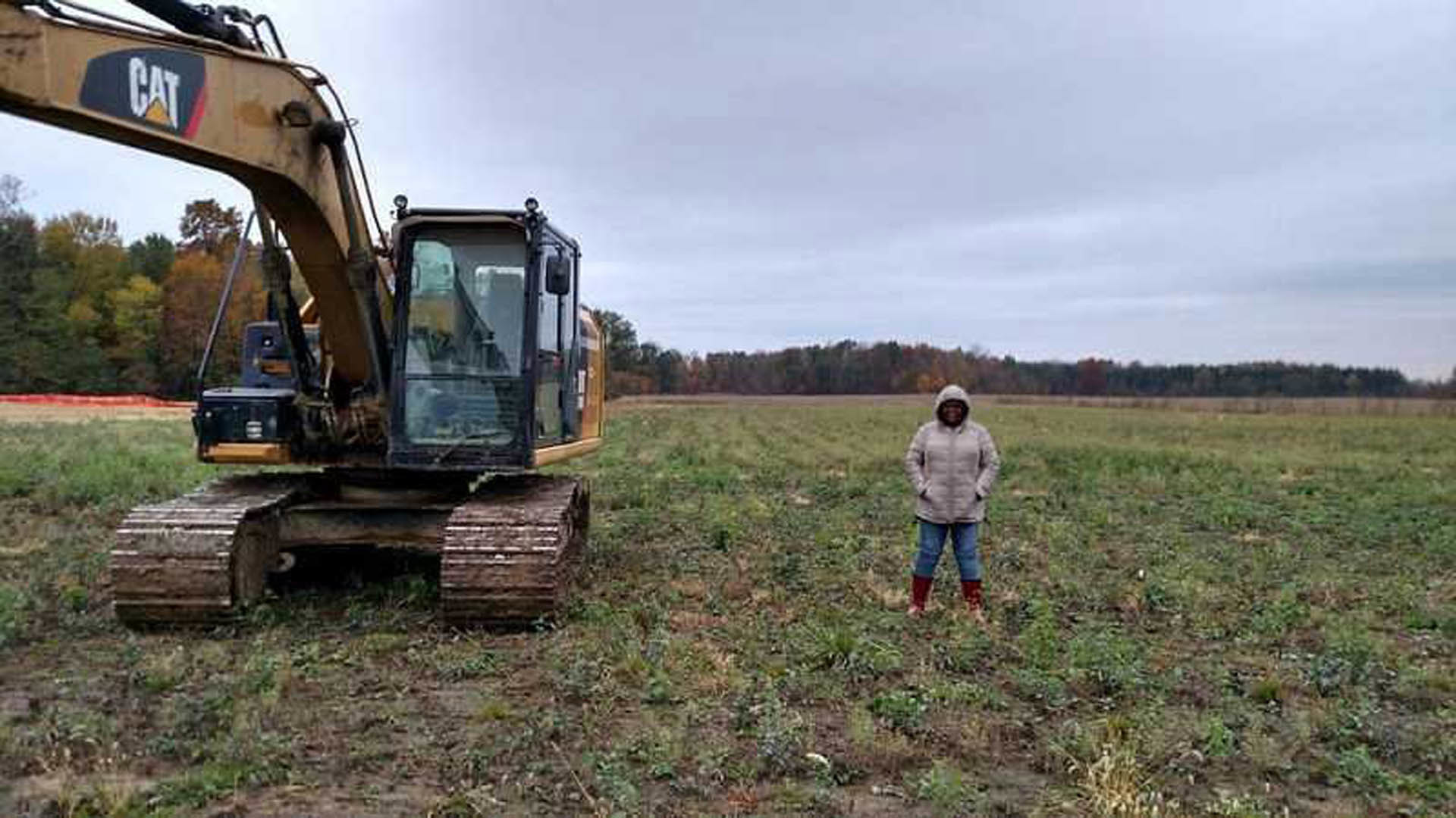 Person in a coat standing on grassy field beside yellow bulldozer, cloudy sky overhead, trees in background