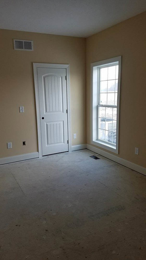 White-painted room featuring a paneled door with a round knob, large window with exterior view and vent, smooth plaster walls with a black trim line, and light-colored flooring.