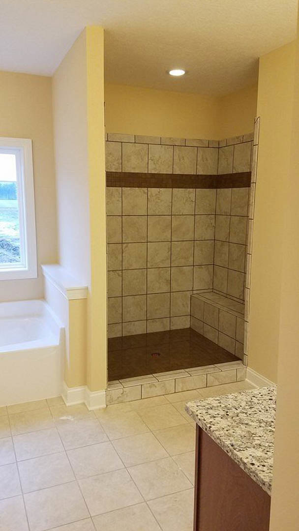 Walk-in shower with glass enclosure, white tile walls, black accent line, brown tile floor with linear drain, quartz countertop, window with frosted glass.