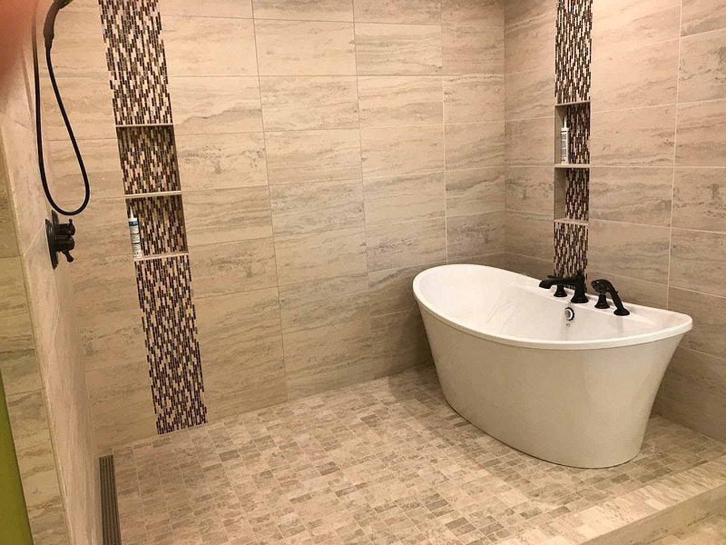 White freestanding bathtub with matte black faucet, gray tile floor, built-in shower shelf holding a bottle, light-colored wall tiles, modern bathroom fixtures