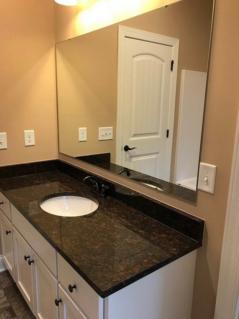 Bathroom with white sink and rectangular mirror above, light switch on wall, white door with black hinges, tile backsplash, and dark cabinetry below countertop