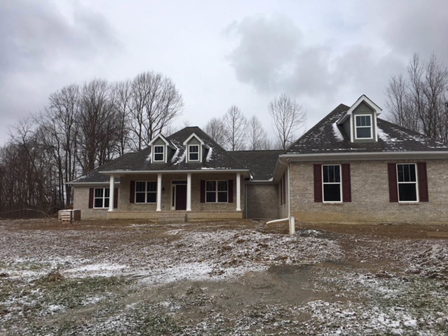 Two-story cottage-style home with white-framed windows, covered porch, and snow-covered yard; trees and cloudy sky in background.