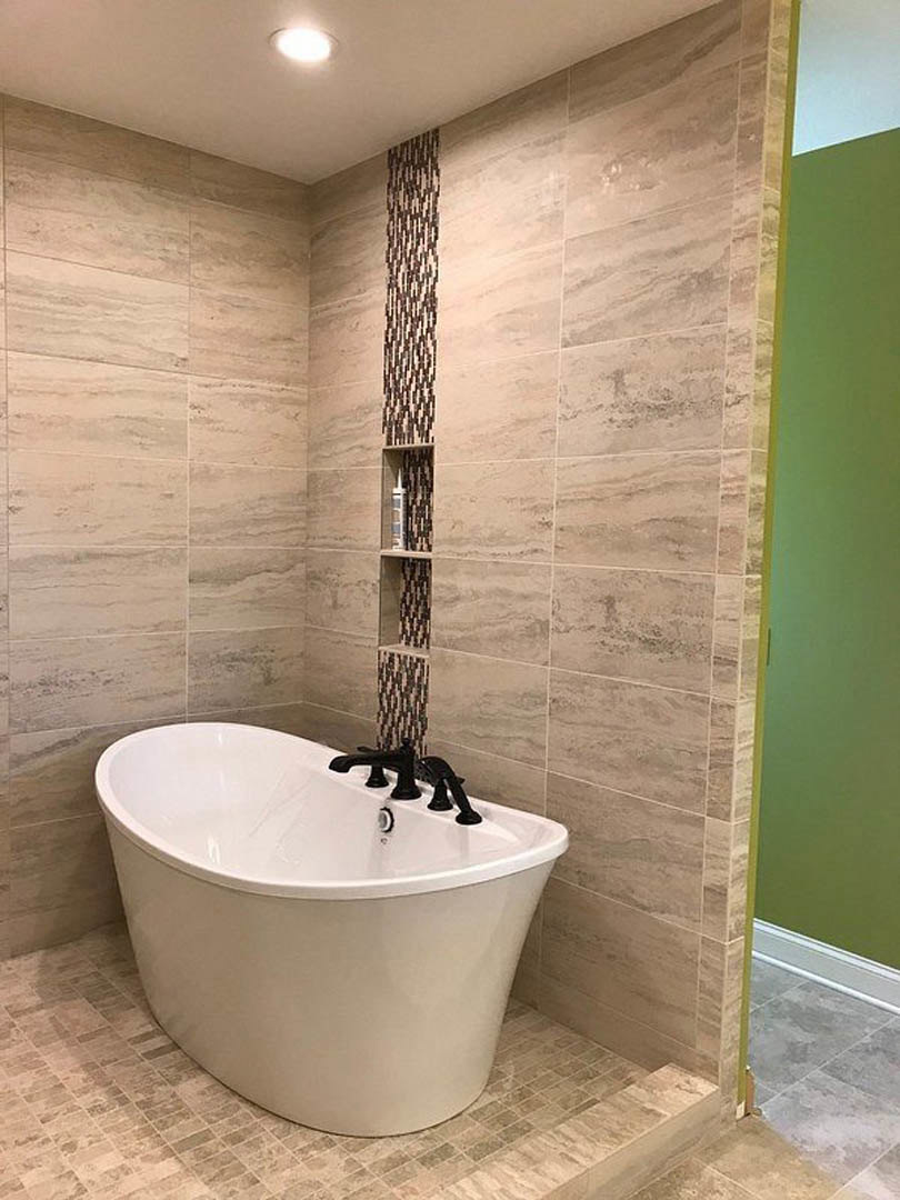 White freestanding bathtub with matte black faucet, green accent wall with tile trim, grey tile flooring bordered in white, ceiling light overhead