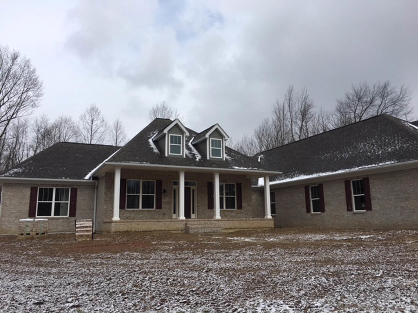 Two-story house with white-framed windows, covered porch, and snow on the roof and ground, surrounded by bare trees and a cloudy sky