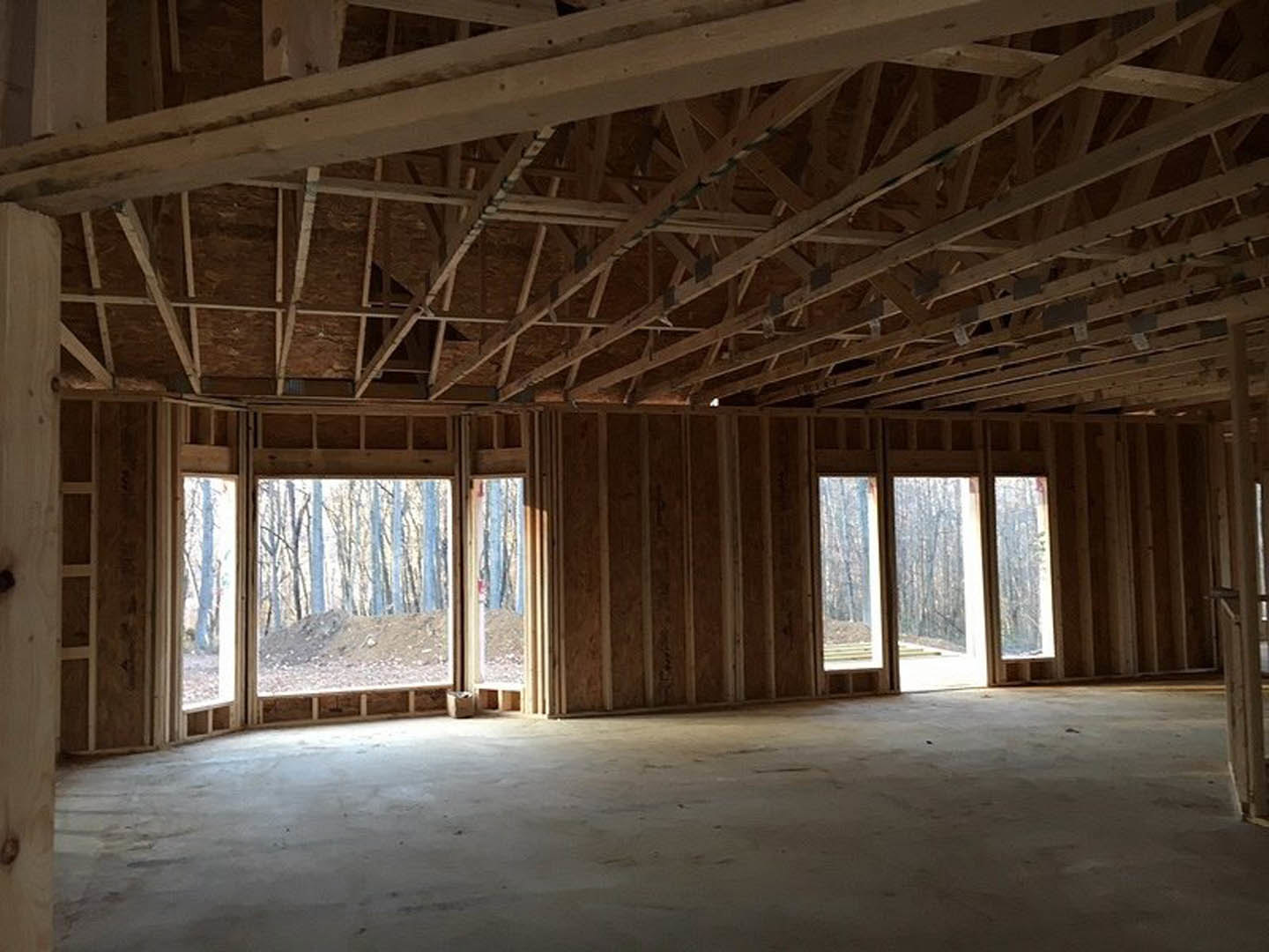 Open room with exposed wooden ceiling beams, large windows, concrete floor, and sunlight streaming in; open door leads to forested landscape visible outside.