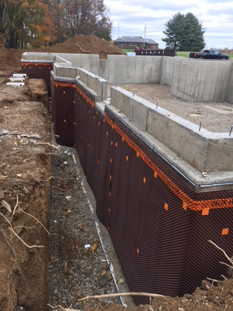 Concrete slab foundation surrounded by dirt and construction materials, with trees and a truck visible in the background under a clear sky.