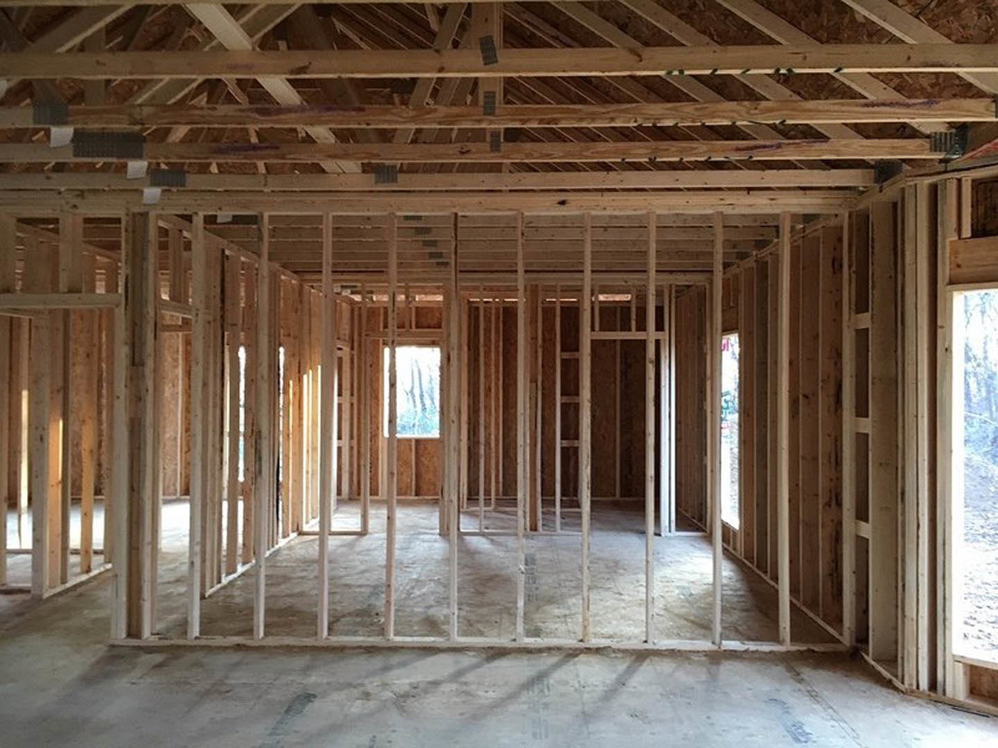 Wood-framed room under construction with exposed ceiling beams, unfinished walls, large window, and concrete floor