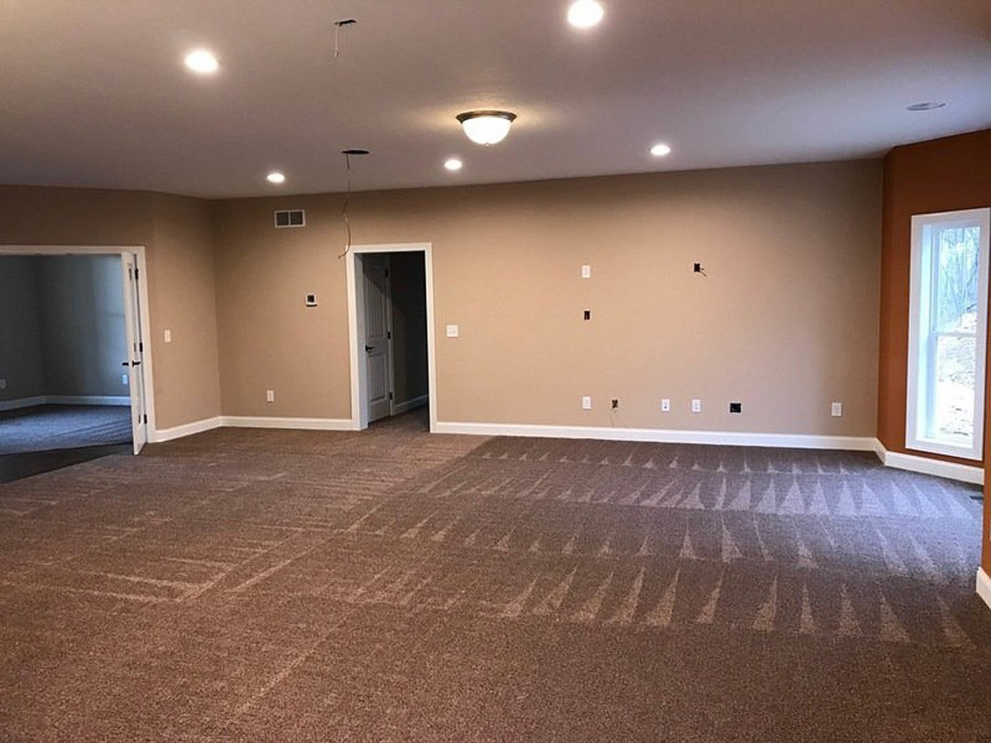 Carpeted room with white walls, recessed ceiling lights, open white door with black handle, large window showing outdoor view, empty space with neutral finishes