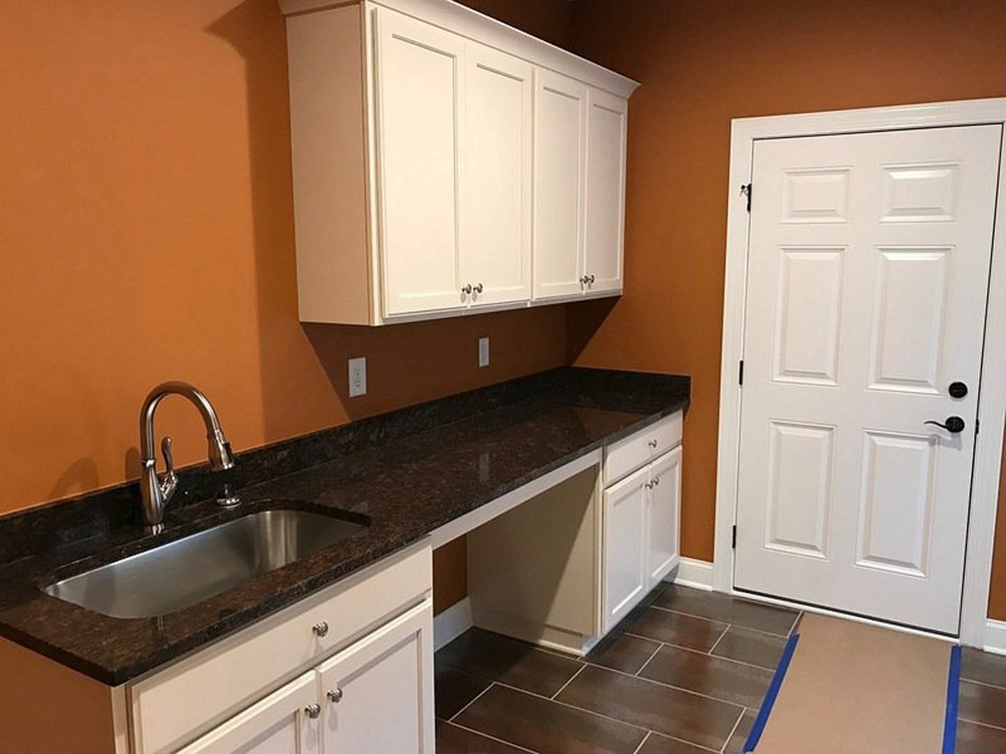 White kitchen cabinets with silver knobs, a stainless steel sink and faucet, blue and white tile flooring, white door with black handles, light-colored countertops
