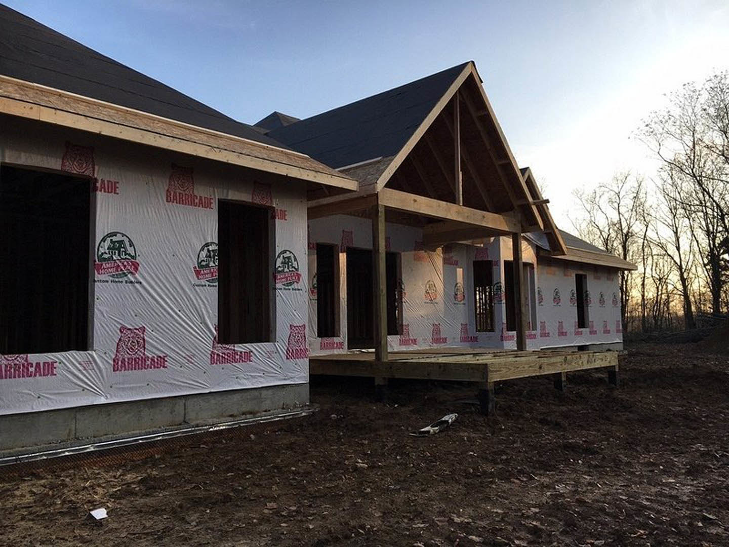 Wood-framed house under construction with covered porch, exposed siding, multiple windows, and unfinished ground surrounded by trees