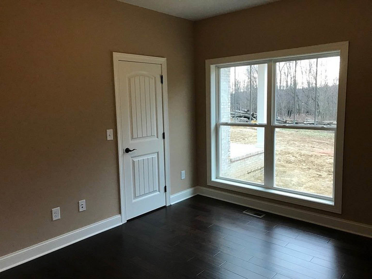 Dark wood flooring with white baseboards, white door with brushed metal handle, large window overlooking green yard, neutral painted walls, light switch near door