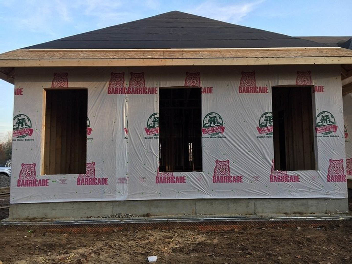 Partially built house with white plastic sheeting covering exterior walls, exposed framing, dark shingle roof with triangular gable, dirt ground in foreground, cloudy sky above