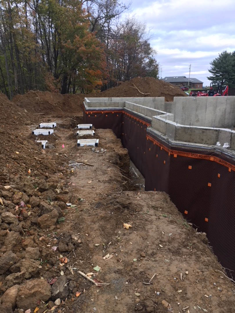 Concrete house foundation surrounded by black tarp at construction site, bordered by trees with green and brown leaves under a cloudy sky