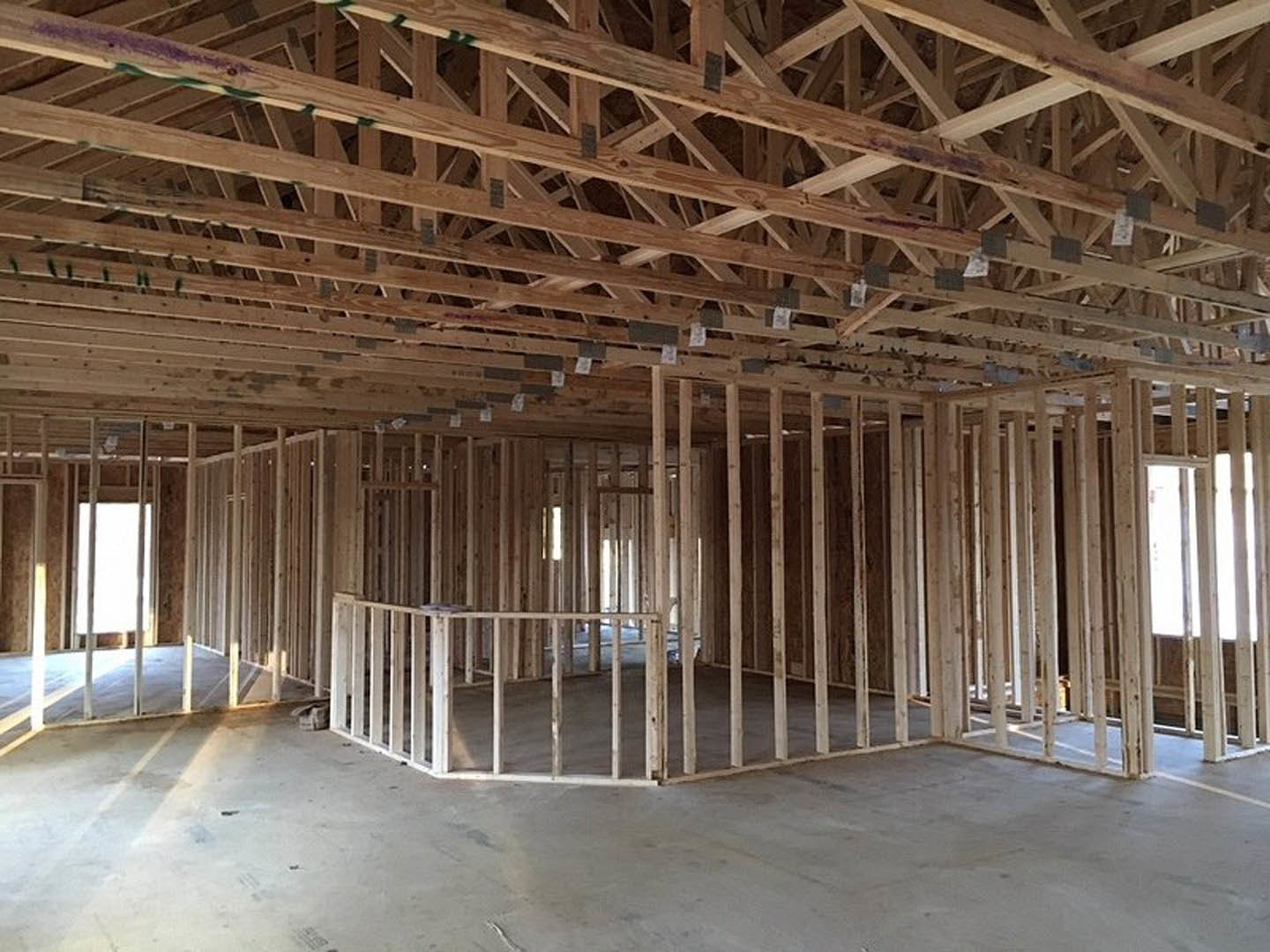 Wood-framed house under construction with exposed beams, unfinished wooden ceiling, concrete floor, and partial wooden railing against a cloudy sky.