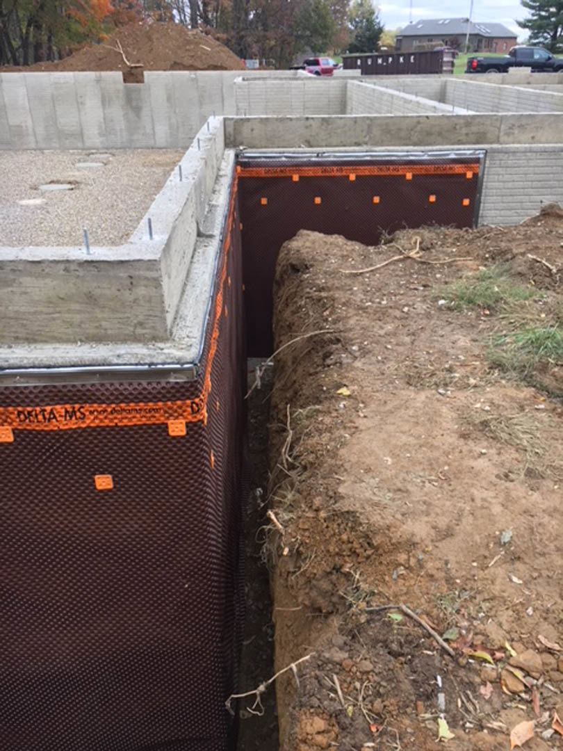 Concrete slab foundation surrounded by dirt, grass, and scattered sticks, with trees and construction materials visible in the background