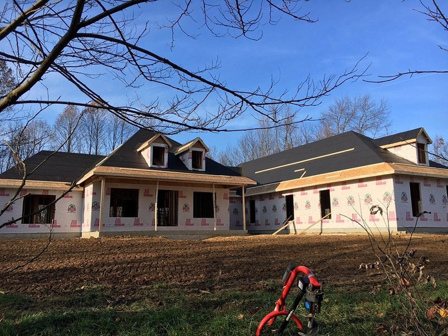 Partially built house with exposed framing, white siding, and unfinished roof; red bicycle parked on grass in front; large windows and door openings visible; leafy tree and blue