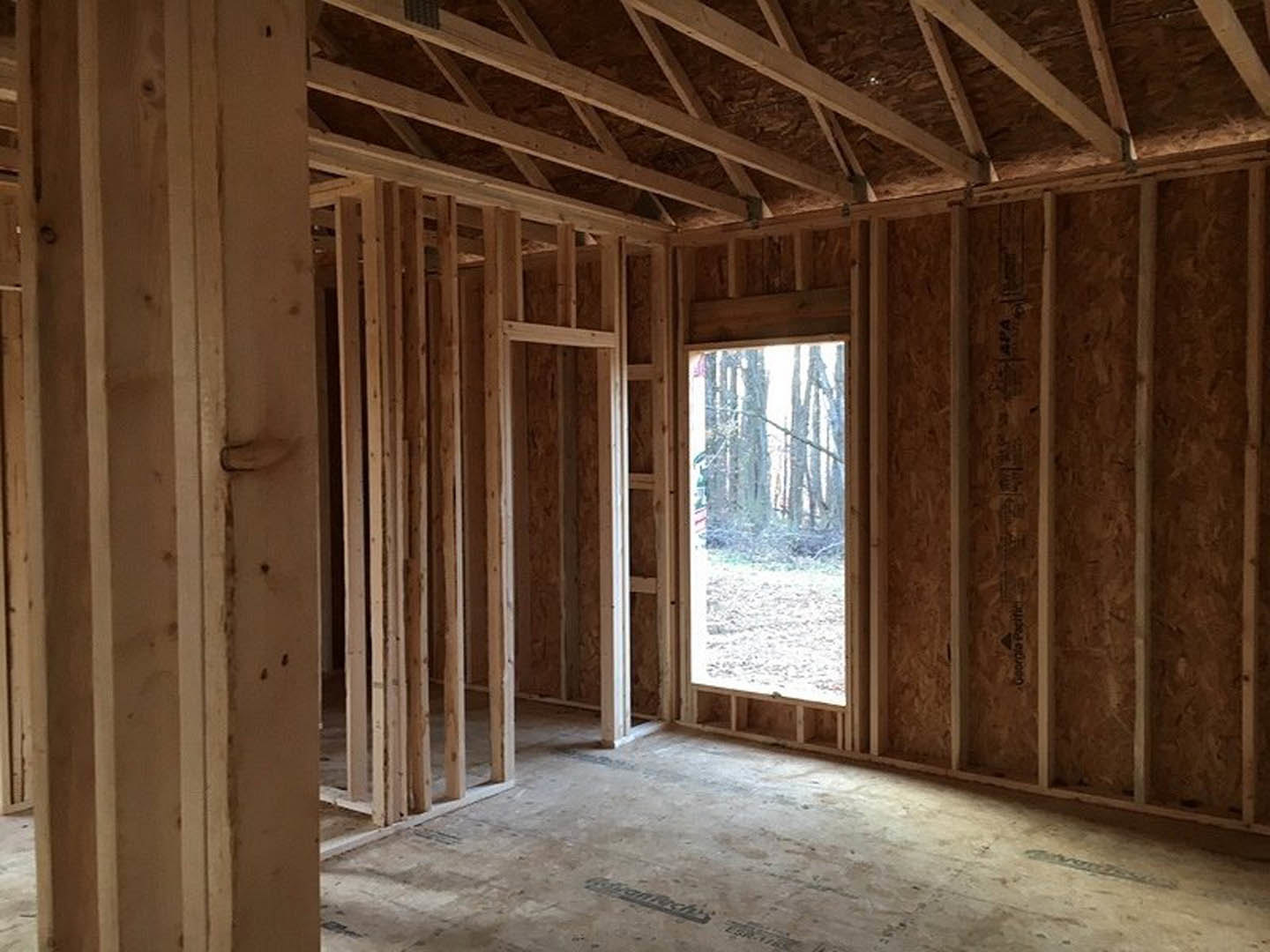 Wood-framed room with exposed beams, large window overlooking snow-covered forest, sunlight illuminating unfinished floor