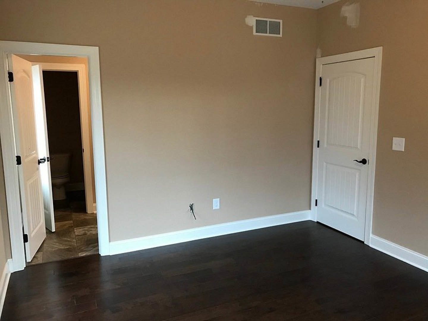 White paneled door with black handle opens to a bathroom featuring a white toilet, dark wood laminate flooring, white baseboards, and a white light switch on plaster wall.