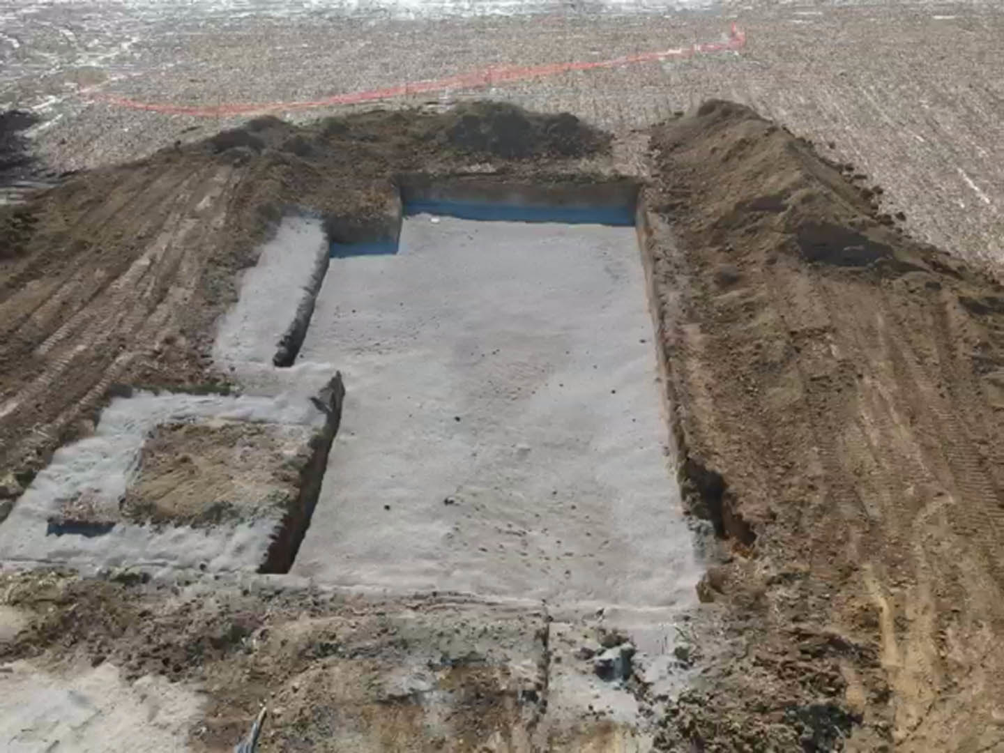 Concrete foundation set in excavated dirt, surrounded by tire tracks and construction materials