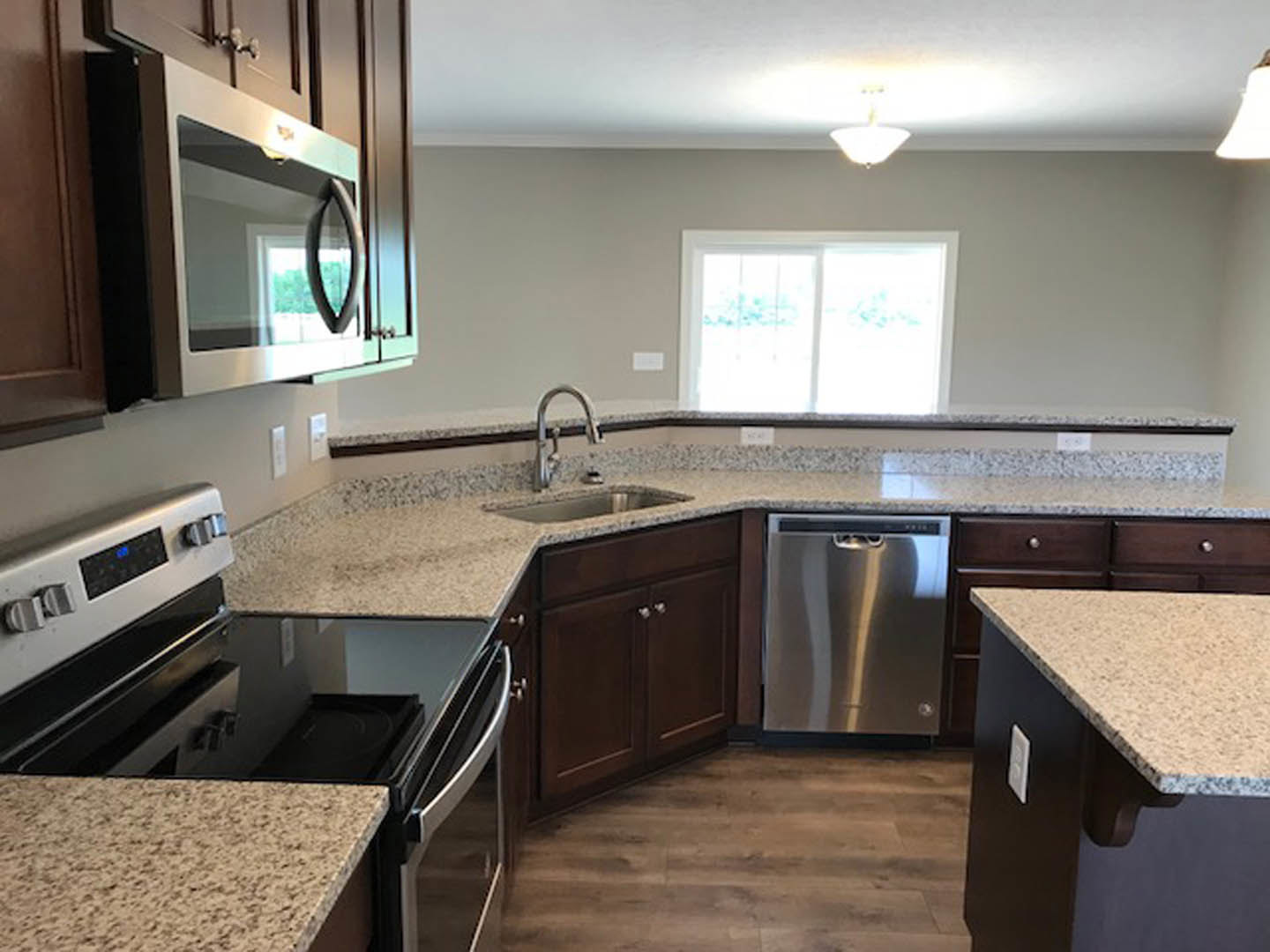 Kitchen with polished granite countertops, dark stovetop, stainless steel microwave with glass door, silver cabinet hardware, white-framed window, and wood cabinetry.