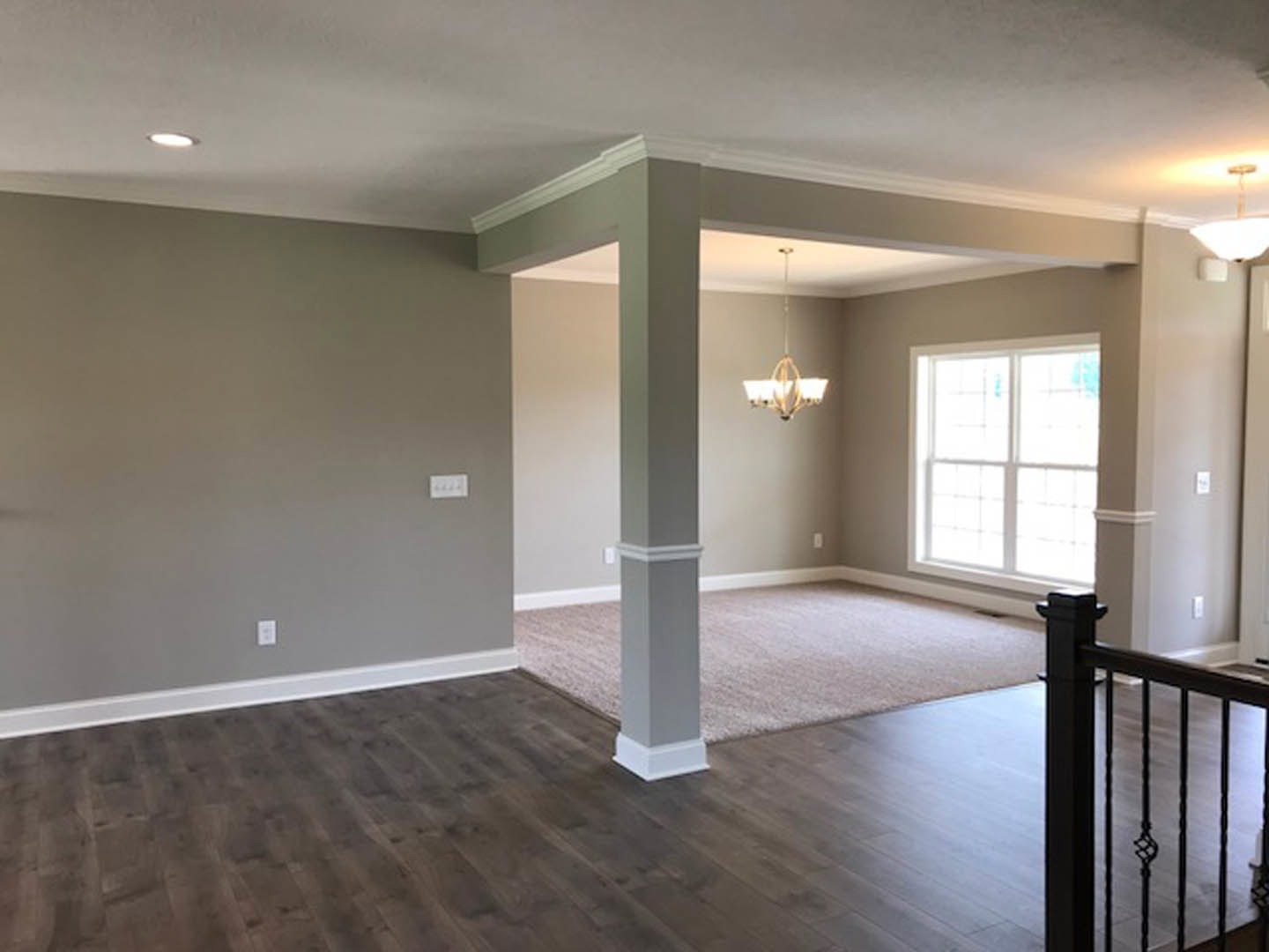 Open living area featuring a modern staircase with black railing, white plaster walls, wood laminate flooring, large window with natural light, and chandelier with white shades.