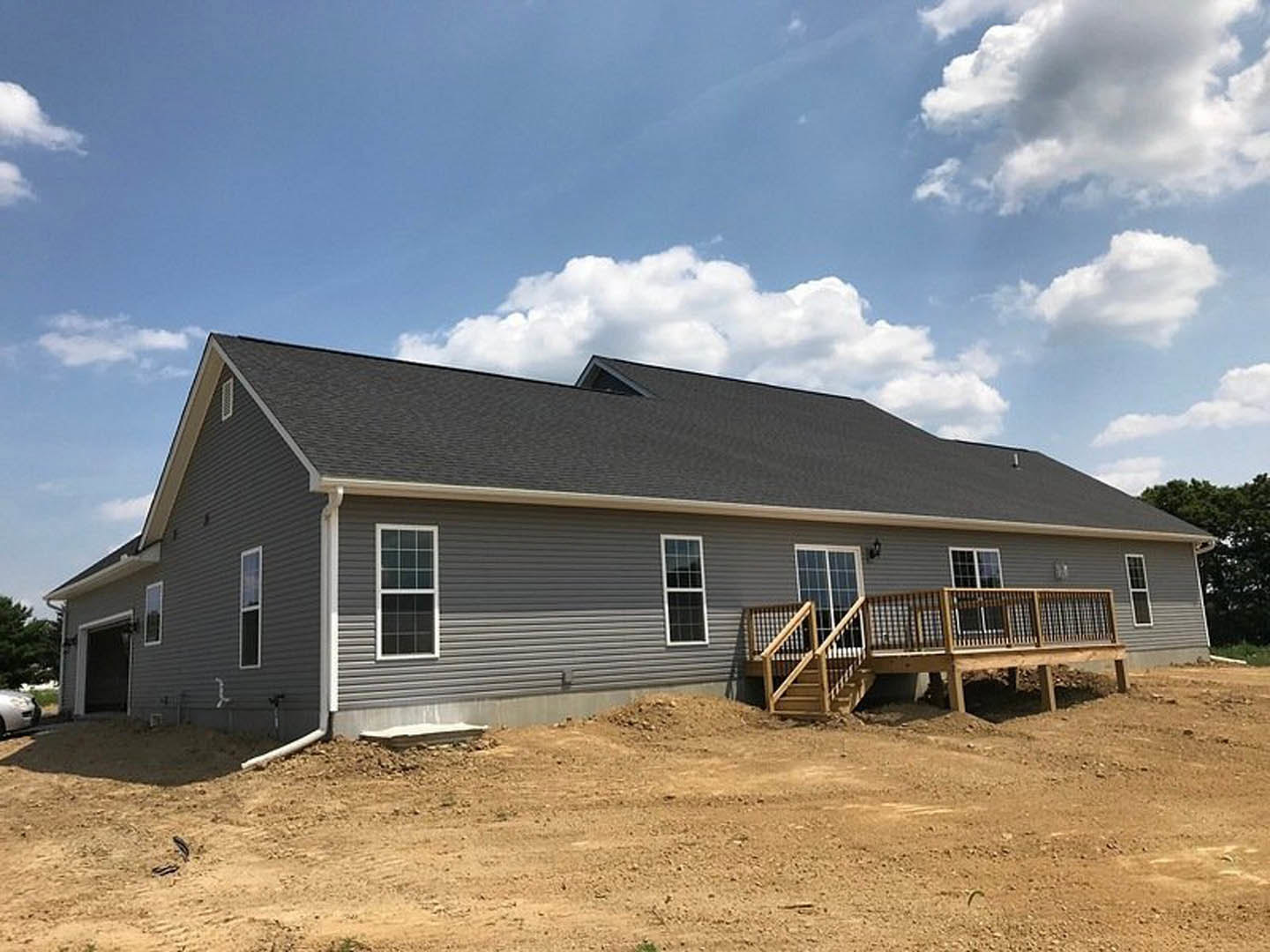 Two-story house under construction featuring a wooden deck with railing, exposed wooden staircase, white-framed windows, and dirt patch in front yard.