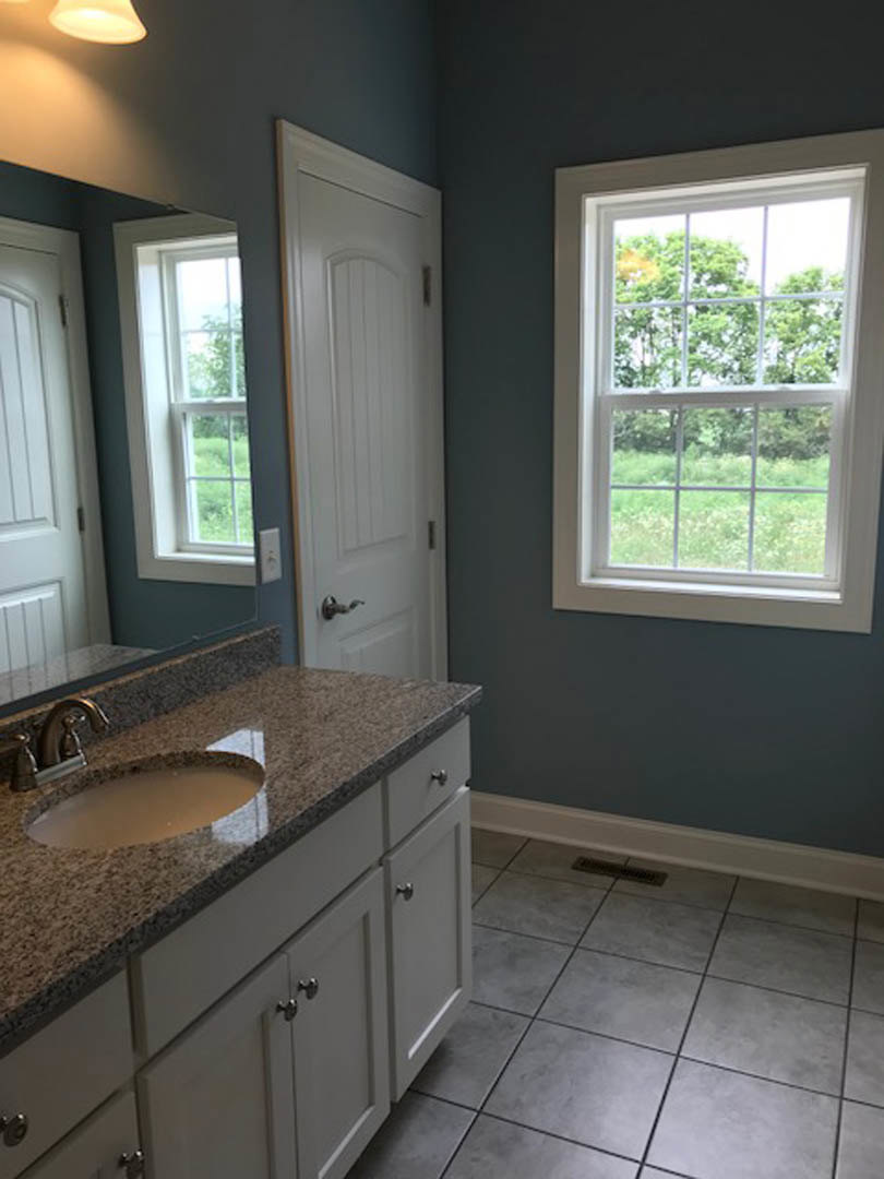 White bathroom with rectangular window, modern chrome faucet, undermount sink set in light stone countertop, white cabinetry, and gray tile floor