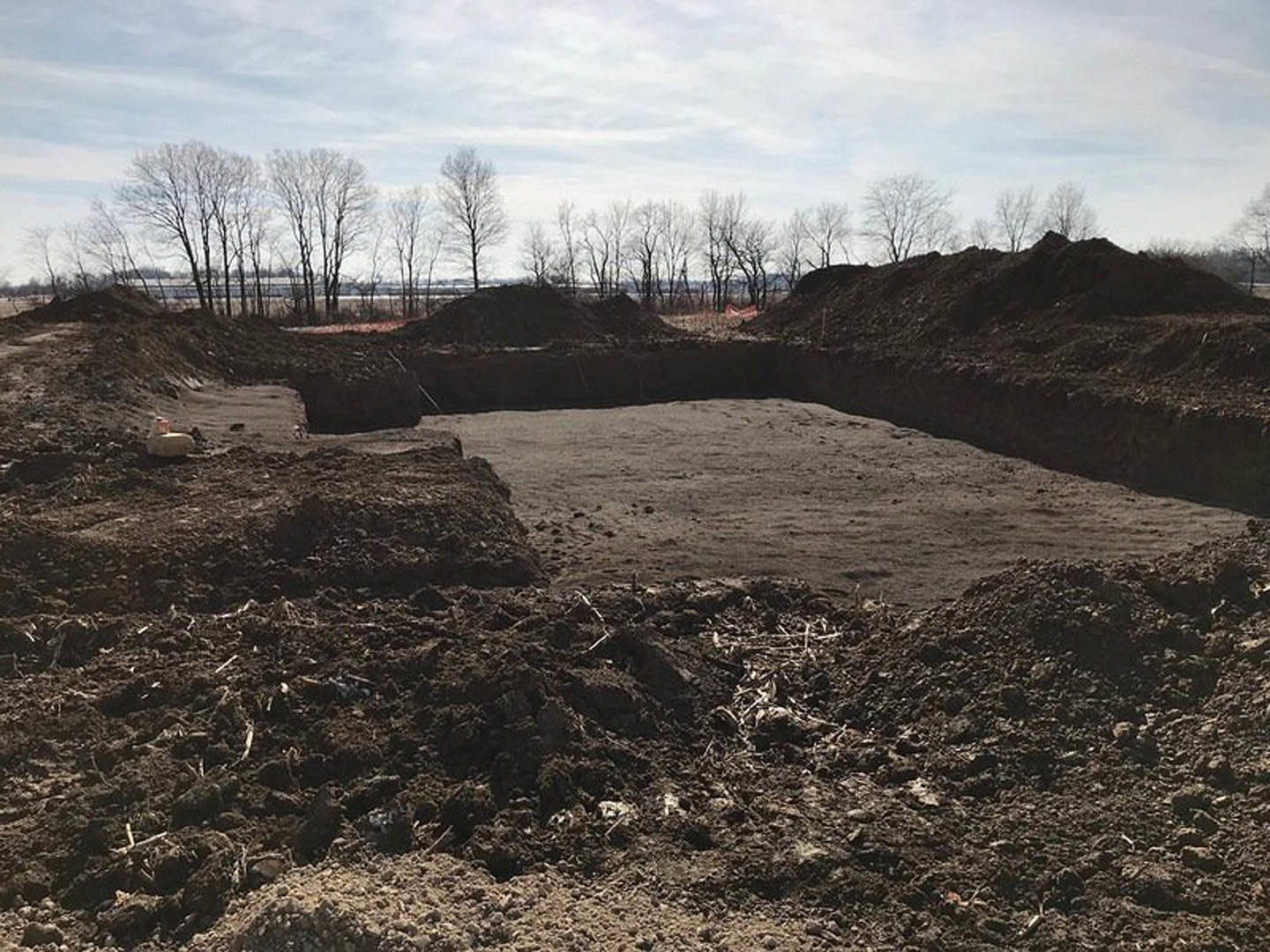 Large mound of loose soil in an open field with leafless trees and cloudy sky in the background