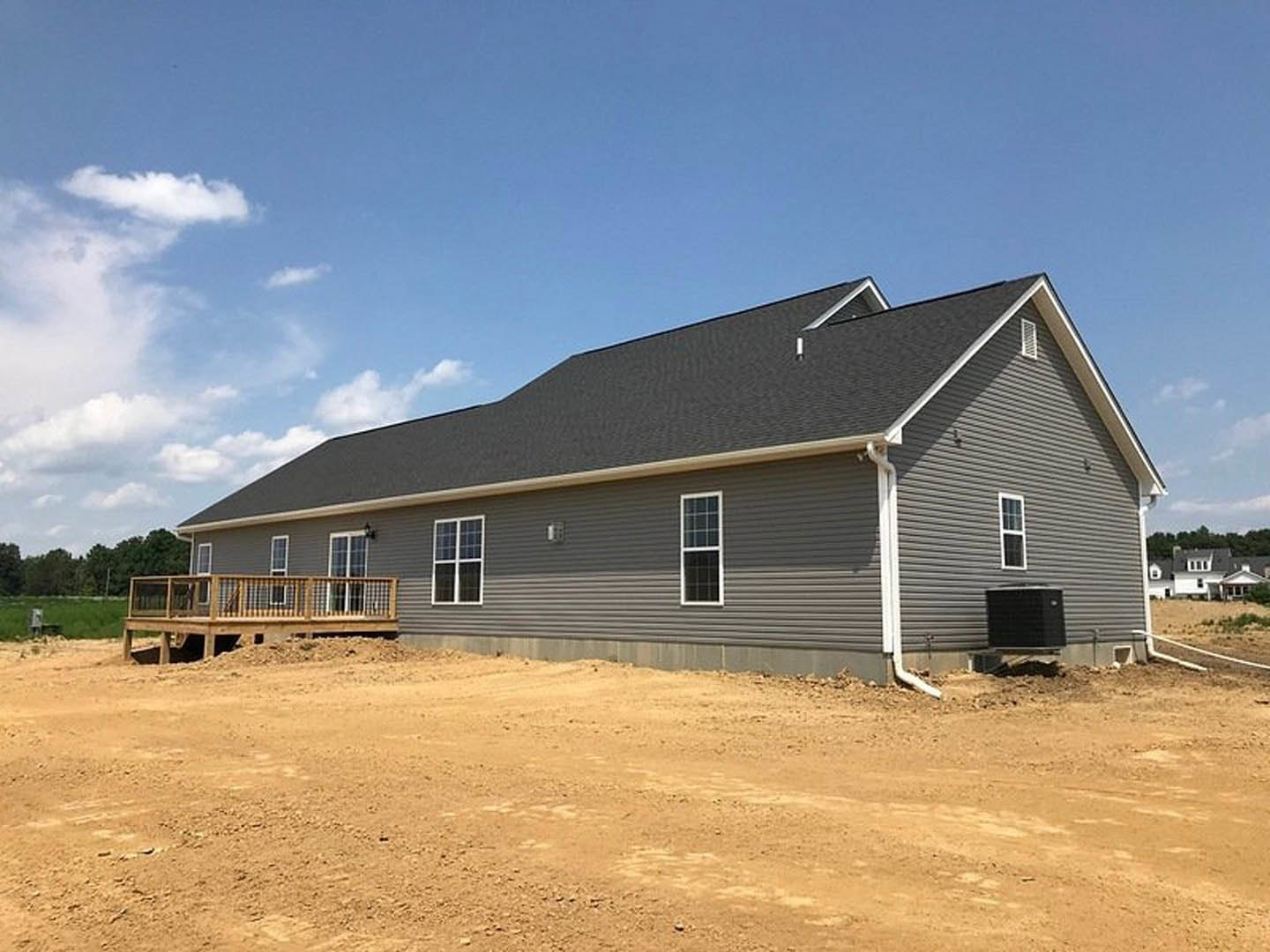 Partially built house with white-framed window, wooden deck and railing, metal retaining wall, and exposed dirt lot under cloudy sky
