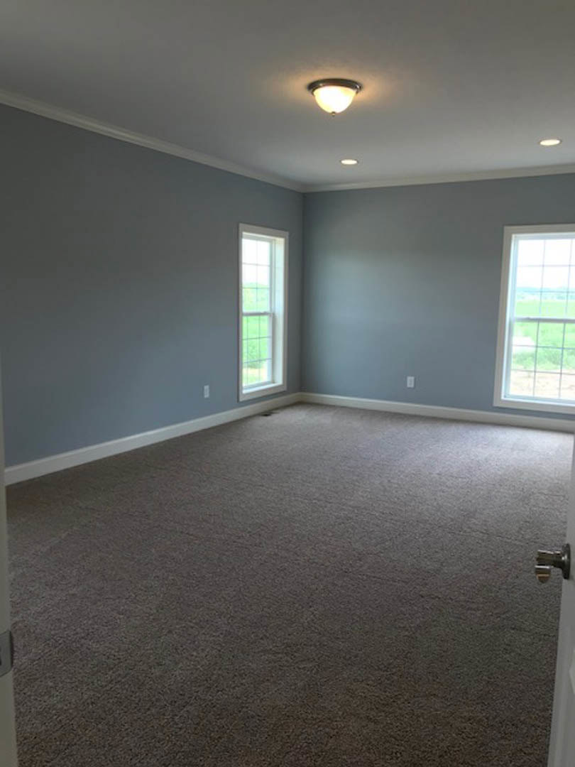 Carpeted room with white walls, ceiling-mounted light fixture, large windows overlooking green grass, and a white door