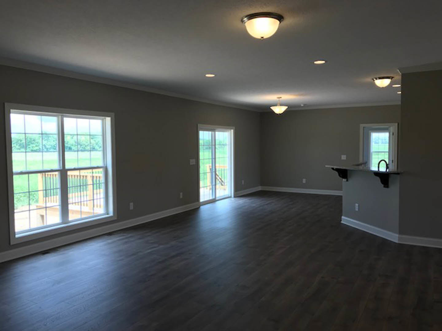 Open kitchen with stainless sink, dark wood flooring, white plaster walls, ceiling light fixture, large windows overlooking deck, grass, and fenced yard