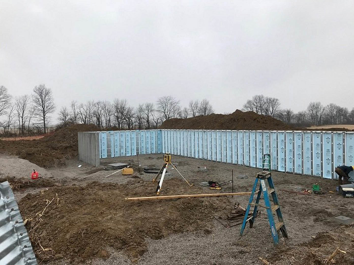 Partially built home foundation surrounded by dirt and soil, blue ladder lying on the ground, metal doors and construction materials visible, trees and cloudy sky in background