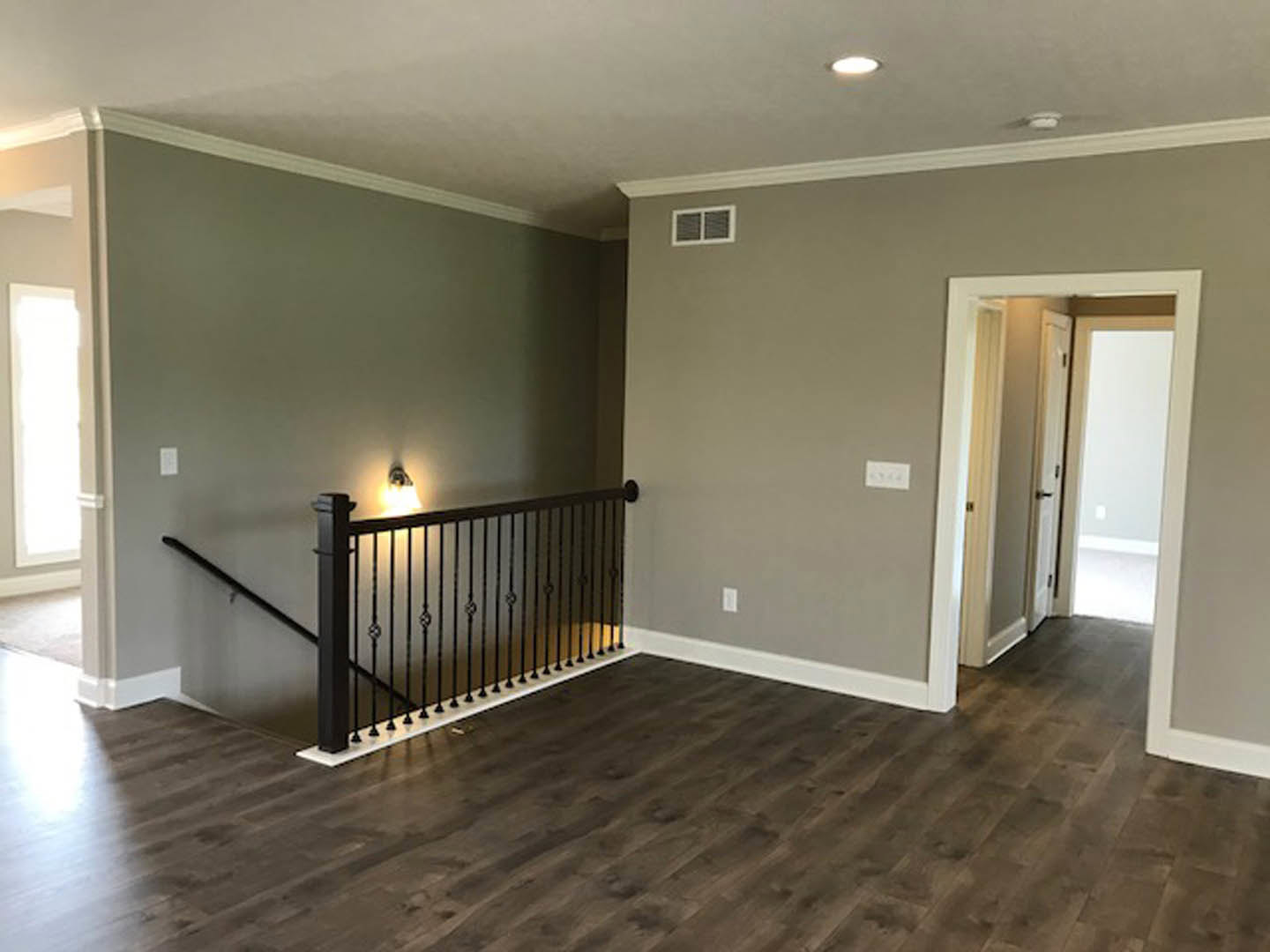 Open room featuring a wood floor, black metal staircase railing, white walls, doorway, and large window with vent.