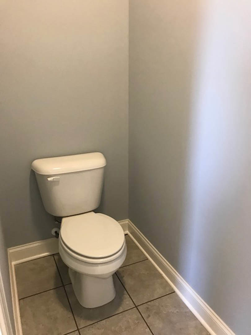 White porcelain toilet with lid closed, chrome flush handle, mounted against light-colored tiled wall, toilet paper roll resting on top of tank, bathroom floor with neutral tile.