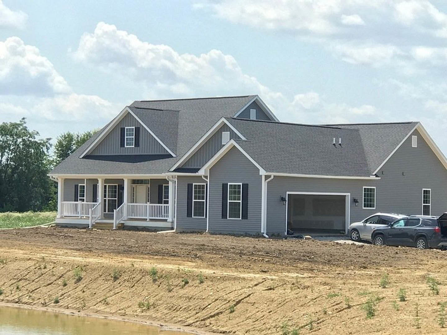 Grey custom home with attached garage, white-framed windows, and a car parked on a dirt driveway; person visible near front window, cloudy sky overhead.