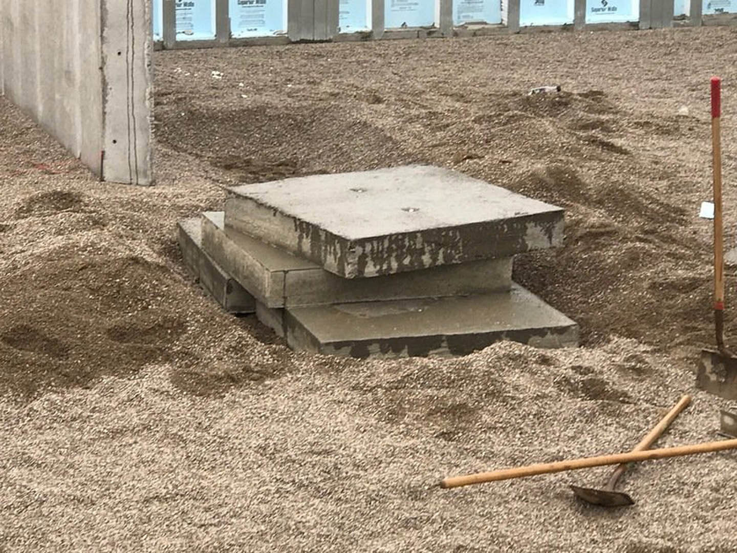 Stacked concrete blocks resting on sandy soil at a construction site, with a shovel nearby and blurred building and wooden post in the background.
