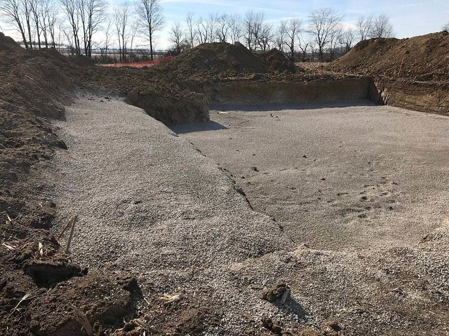 Excavated dirt pit with exposed soil and gravel, metal pole near a leafless tree, cloudy sky above, surrounded by grassy field and distant trees