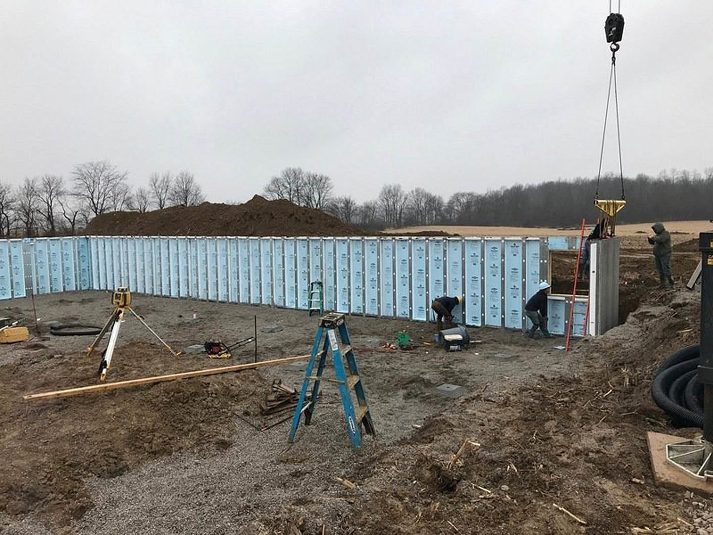 Crane hoisting prefabricated wall panel at residential construction site, foundation visible, workers observing, hillside with trees in background