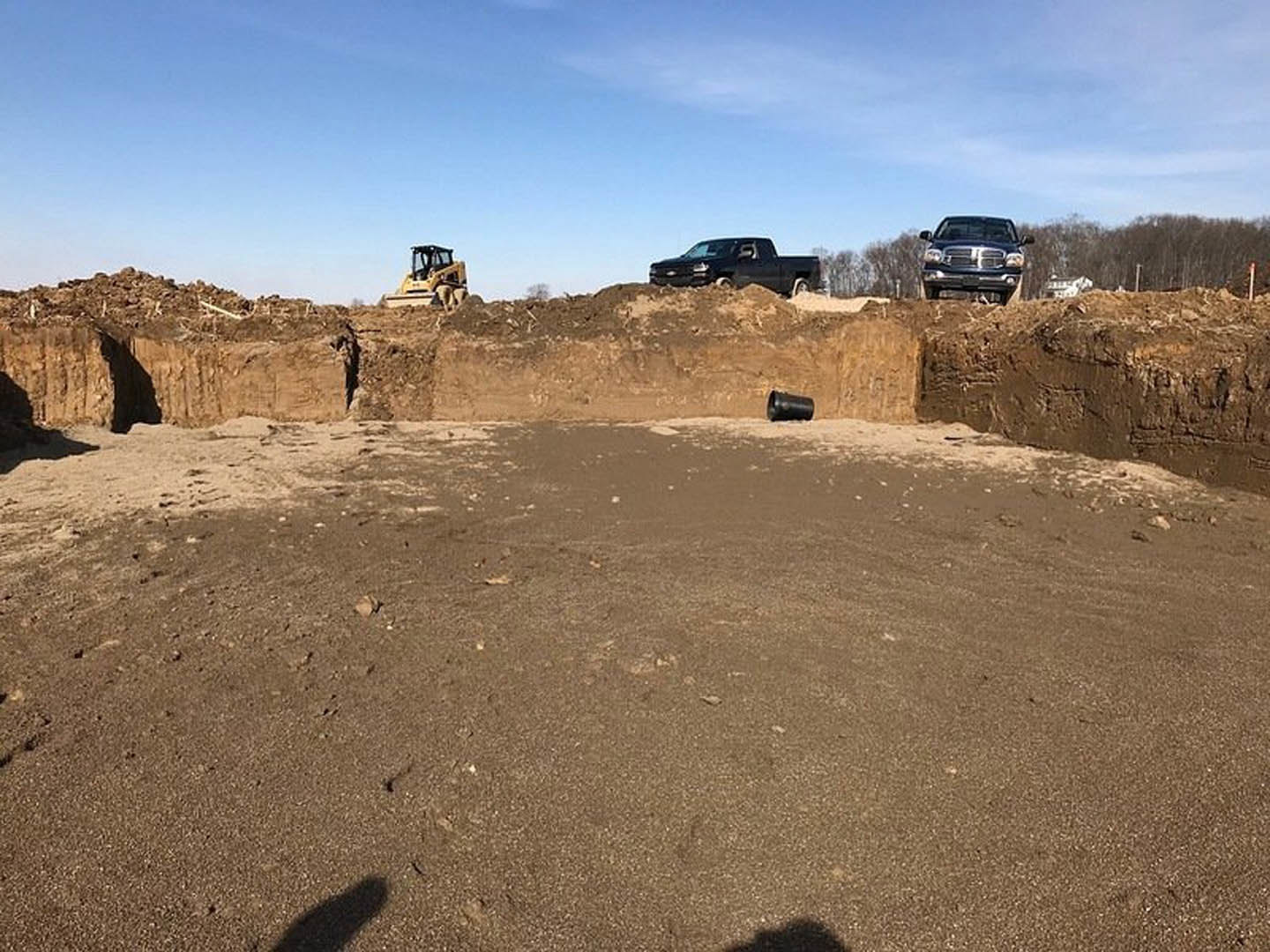 Dirt construction site with parked trucks and cars, cloudy sky overhead, soil and scattered equipment visible