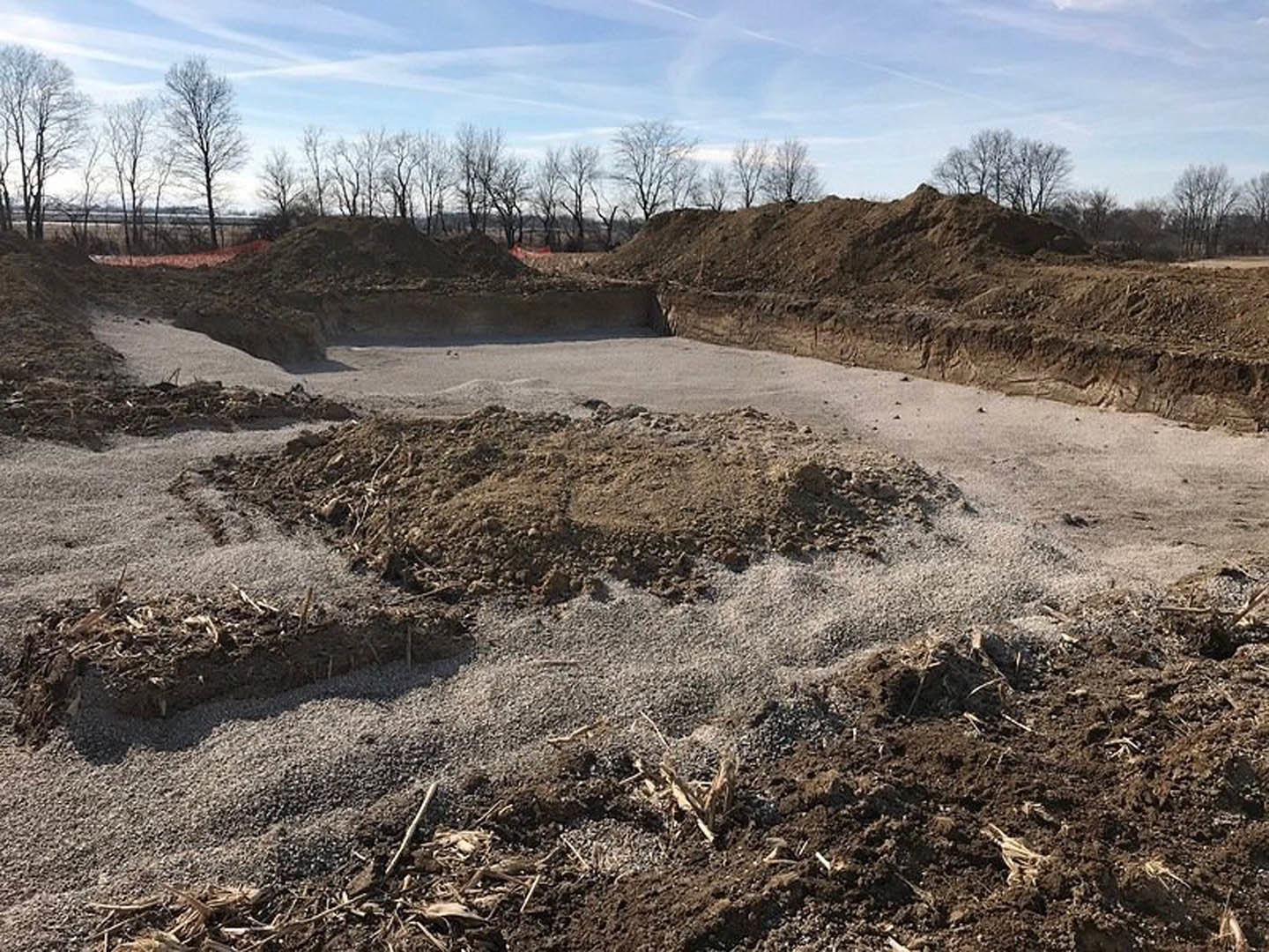 Large dirt mound in foreground with scattered leafless trees and cloudy sky in background