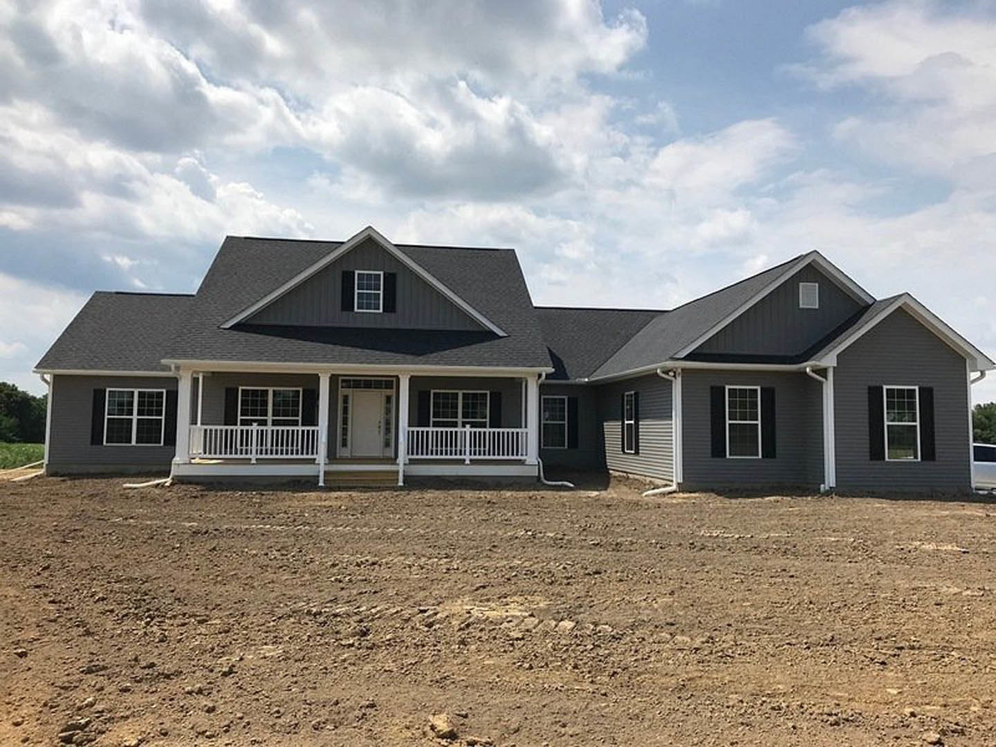 Two-story house under construction with white porch, white-framed windows, glass-paneled front door, dirt yard with tire tracks, gray shingle roof, and cloudy sky.