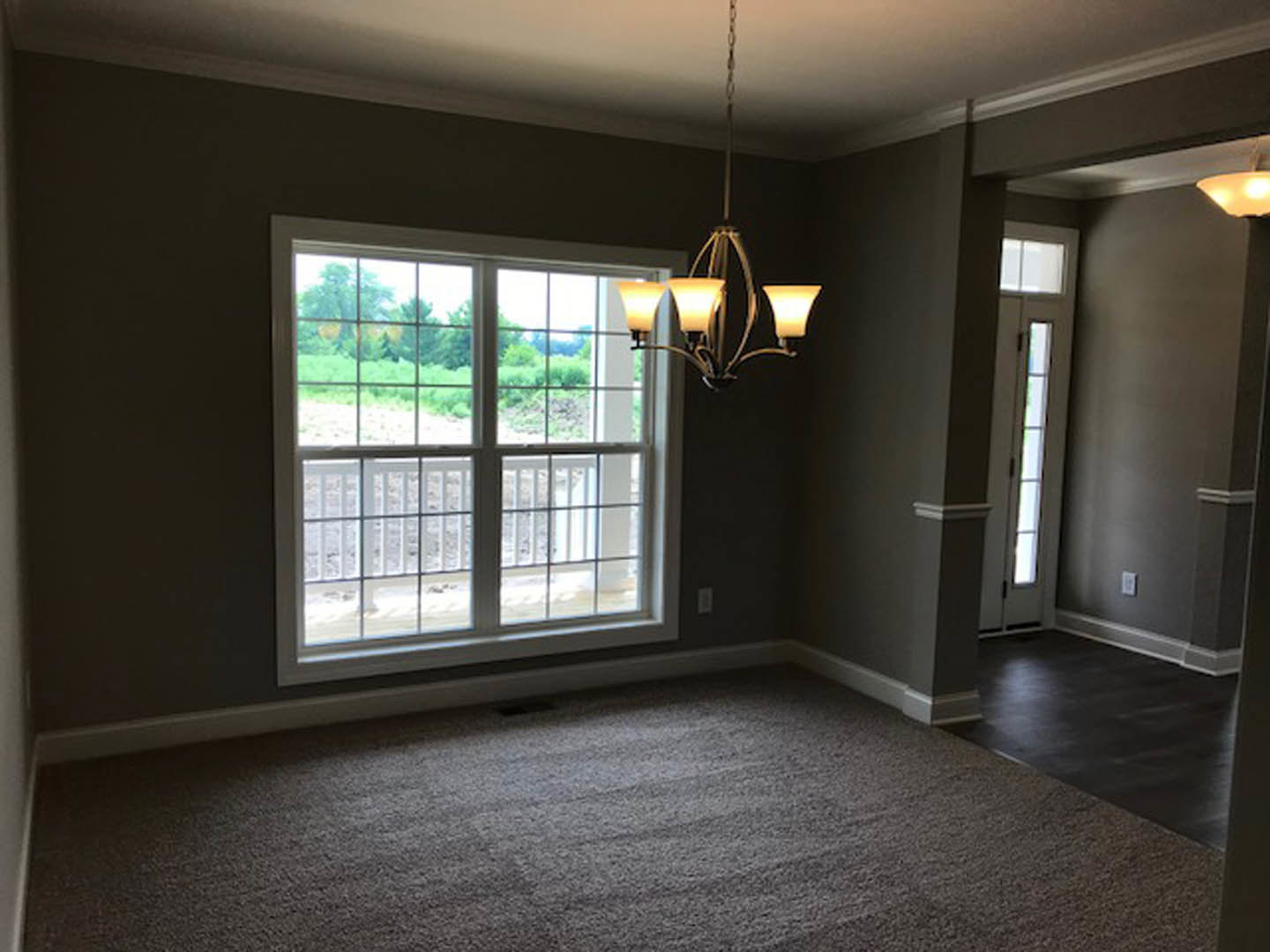 Chandelier hanging from white ceiling, large window with daylight, hardwood flooring, neutral walls, interior door visible
